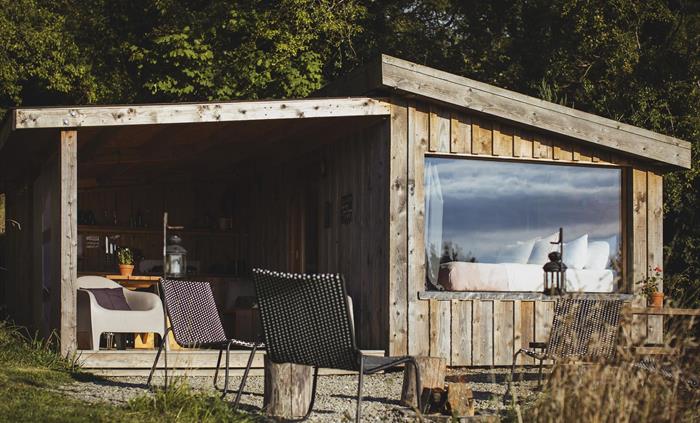 Wood cabin set amongst woodland.  There is a covered outdoor camp kitchen and a firepit to the foreground. Inside is a bed made up with a view out of